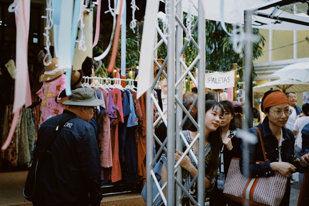 A bustling outdoor market scene with several people browsing clothes on display racks. Brightly colored garments hang alongside streamers. A canopy provides shade, while a sign reading 'PALETAS' is visible in the background. Shoppers are engaged in conversation and appear to be enjoying the lively atmosphere.