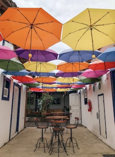 A narrow outdoor corridor with colorful umbrellas hanging overhead, creating a vibrant canopy. Below are wooden tables and high stools, set against white walls with blue trim. Potted plants and artwork hang on the walls, enhancing the cozy atmosphere.