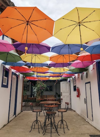 A narrow outdoor corridor with colorful umbrellas hanging overhead, creating a vibrant canopy. Below are wooden tables and high stools, set against white walls with blue trim. Potted plants and artwork hang on the walls, enhancing the cozy atmosphere.