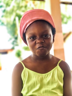 A young child with a playful expression is wearing a bright pink headscarf and a lime green sleeveless top. The background is softly blurred, suggesting an outdoor setting with greenery.
