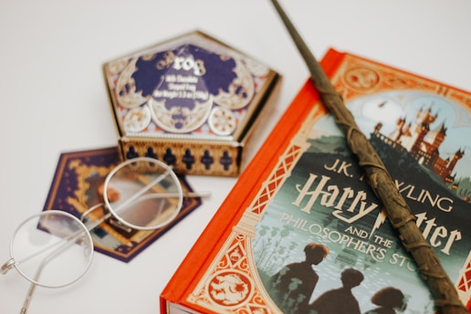 A collection of fantasy-themed items including a Harry Potter book with a detailed cover, a wand, a pair of round spectacles, and a decorative box labeled 'Frog'. These items are arranged on a white surface, showcasing a magical and whimsical theme.