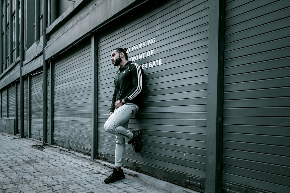 man in black jacket and white pants leaning on gray wall