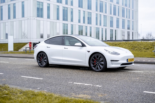 A white electric car is parked on a paved road in front of a modern glass and steel office building. The building features large vertical windows and is bordered by neatly trimmed grass. The car, with its sleek and aerodynamic design, features dark alloy wheels with red brake calipers, adding a sporty accent to its appearance.