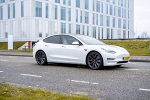 A white electric car is parked on a paved road in front of a modern glass and steel office building. The building features large vertical windows and is bordered by neatly trimmed grass. The car, with its sleek and aerodynamic design, features dark alloy wheels with red brake calipers, adding a sporty accent to its appearance.