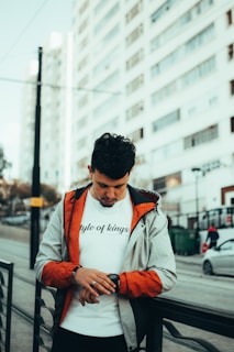 A man wearing a casual jacket and a white t-shirt with 'style of kings' printed on it is standing in an urban setting. He is looking down at a smartwatch on his wrist. The background shows tall residential buildings and a street setting with cars and pedestrians.