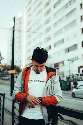 A man wearing a casual jacket and a white t-shirt with 'style of kings' printed on it is standing in an urban setting. He is looking down at a smartwatch on his wrist. The background shows tall residential buildings and a street setting with cars and pedestrians.