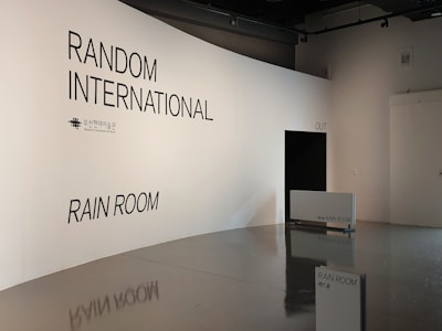 An art exhibit space with a sleek, modern design. The walls are white, displaying the text 'RANDOM INTERNATIONAL' and 'RAIN ROOM' prominently. There is a black opening labeled 'OUT' and signs indicating the entrance to the Rain Room.