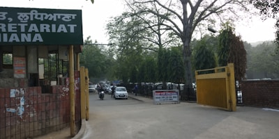 The image depicts the entrance to a government or administrative building with signs in different languages. Tall trees line the street, and several vehicles, including cars and a motorcycle, are visible on the road. A sign indicating a checkpoint is positioned near a yellow barrier gate.