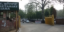 The image depicts the entrance to a government or administrative building with signs in different languages. Tall trees line the street, and several vehicles, including cars and a motorcycle, are visible on the road. A sign indicating a checkpoint is positioned near a yellow barrier gate.