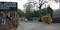 The image depicts the entrance to a government or administrative building with signs in different languages. Tall trees line the street, and several vehicles, including cars and a motorcycle, are visible on the road. A sign indicating a checkpoint is positioned near a yellow barrier gate.