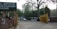 The image depicts the entrance to a government or administrative building with signs in different languages. Tall trees line the street, and several vehicles, including cars and a motorcycle, are visible on the road. A sign indicating a checkpoint is positioned near a yellow barrier gate.
