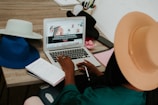 A person wearing a beige hat is seated at a desk, using a laptop computer. Several other hats in blue, black, and off-white are placed on the desk. A notebook with handwriting and a pen are beside the laptop. The background includes a whiteboard with some sketches or notes.