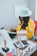 A person wearing a light-colored hat and a yellow floral shirt is sitting at a desk writing in a notebook. The desk is cluttered with a closed laptop, a package with a shipping label, a small trophy, some books, and various office supplies. A whiteboard with cursive writing is partially visible in the background.
