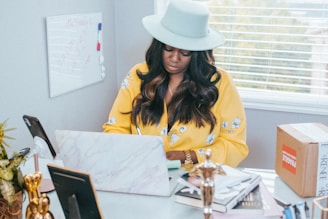 A woman wearing a light blue hat and a yellow floral shirt is writing or taking notes at a desk. The room is bright with natural light coming through the blinds. A laptop with a marble finish is on the desk, along with various items such as a cardboard box labeled 'Fragile,' a smartphone on a stand, and several books and papers. A whiteboard with writing is mounted on the wall.