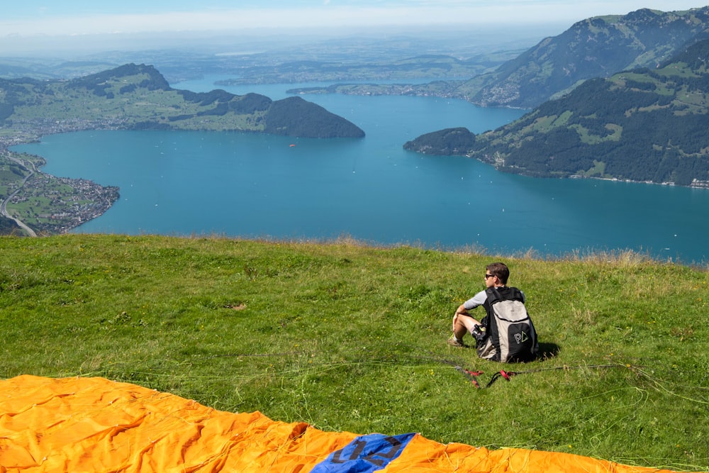 homme en chemise noire et blanche assis sur un champ d’herbe verte près du lac pendant la journée