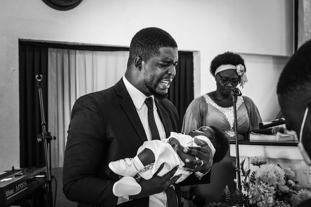man in black suit jacket holding baby in grayscale photography