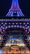 A night view of a tower illuminated with purple and blue lights. At the base, a sign with 'Parisian' written on it in bright red letters is visible, along with decorative golden statues and a fountain. The tower appears to be an architectural structure similar to the Eiffel Tower.
