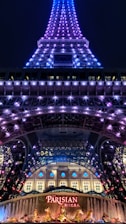 A night view of a tower illuminated with purple and blue lights. At the base, a sign with 'Parisian' written on it in bright red letters is visible, along with decorative golden statues and a fountain. The tower appears to be an architectural structure similar to the Eiffel Tower.