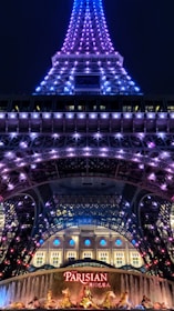 A night view of a tower illuminated with purple and blue lights. At the base, a sign with 'Parisian' written on it in bright red letters is visible, along with decorative golden statues and a fountain. The tower appears to be an architectural structure similar to the Eiffel Tower.