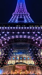 A night view of a tower illuminated with purple and blue lights. At the base, a sign with 'Parisian' written on it in bright red letters is visible, along with decorative golden statues and a fountain. The tower appears to be an architectural structure similar to the Eiffel Tower.