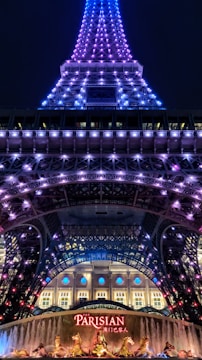 A night view of a tower illuminated with purple and blue lights. At the base, a sign with 'Parisian' written on it in bright red letters is visible, along with decorative golden statues and a fountain. The tower appears to be an architectural structure similar to the Eiffel Tower.