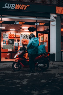 A person wearing a helmet and a teal jacket is standing next to a red scooter with a turquoise delivery bag, presumably for food delivery, in front of a Subway restaurant. The setting appears to be at night or dusk, with the restaurant lights illuminating the scene.