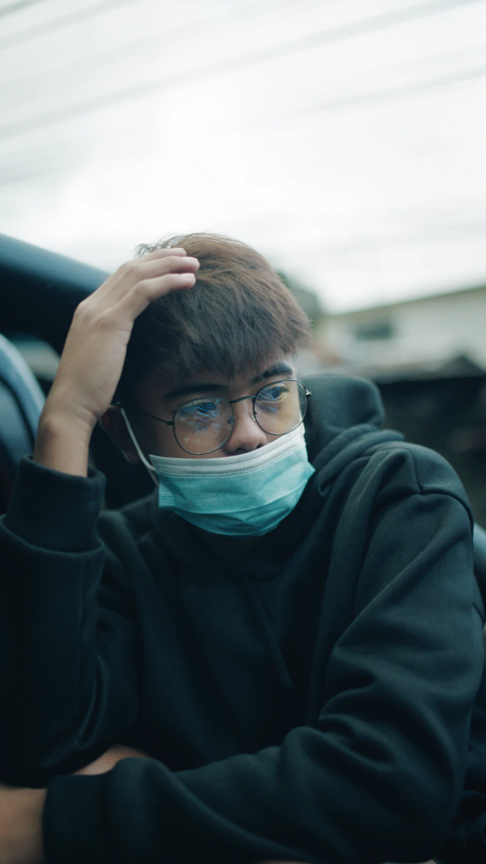 The image shows a young man wearing glasses and a face mask, sitting inside a car with his head resting on his hand. The car is parked against a backdrop of a building, suggesting an urban setting.