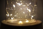 A decorative glass jar containing warmly glowing fairy lights arranged in delicate loops. The golden-hued lights create a cozy ambiance with a soft, diffused glow.