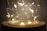 A decorative glass jar containing warmly glowing fairy lights arranged in delicate loops. The golden-hued lights create a cozy ambiance with a soft, diffused glow.