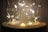 A decorative glass jar containing warmly glowing fairy lights arranged in delicate loops. The golden-hued lights create a cozy ambiance with a soft, diffused glow.