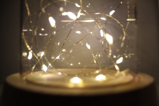 A decorative glass jar containing warmly glowing fairy lights arranged in delicate loops. The golden-hued lights create a cozy ambiance with a soft, diffused glow.