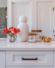 A white countertop features a decorative ceramic vase and a clear glass bowl with vibrant red roses. Next to them is a set of jars with wooden lids and a golden leaf-shaped decor piece. The backdrop includes paneled white cabinets.