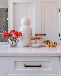 A white countertop features a decorative ceramic vase and a clear glass bowl with vibrant red roses. Next to them is a set of jars with wooden lids and a golden leaf-shaped decor piece. The backdrop includes paneled white cabinets.