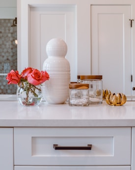 A white countertop features a decorative ceramic vase and a clear glass bowl with vibrant red roses. Next to them is a set of jars with wooden lids and a golden leaf-shaped decor piece. The backdrop includes paneled white cabinets.