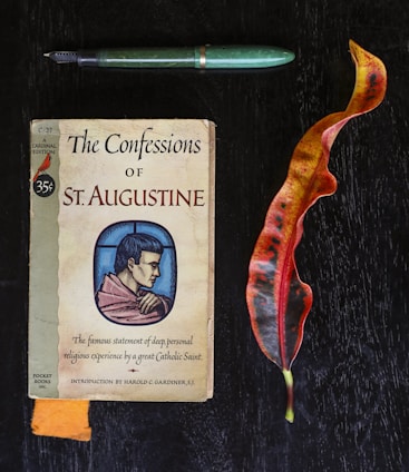 A vintage book titled 'The Confessions of St. Augustine' is placed on a dark wooden surface. Accompanying the book are a green fountain pen and a colorful, elongated leaf with shades of orange and red.