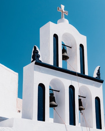 A white stucco building with arches and three large bells hangs against a clear blue sky. The architecture is characteristic of a Greek Orthodox church, featuring clean lines and a cross at the top.