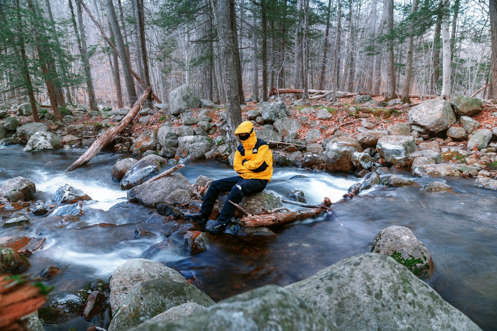 homme en veste jaune et noire assis sur le rocher dans la rivière pendant la journée