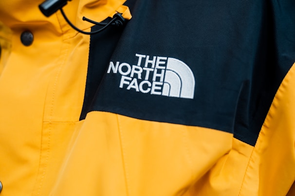 A close-up of a jacket featuring a prominent logo of 'The North Face' embroidered in white on a contrasting black section of fabric. The majority of the jacket is bright yellow with details such as buttons and stitching visible.