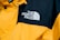 A close-up of a jacket featuring a prominent logo of 'The North Face' embroidered in white on a contrasting black section of fabric. The majority of the jacket is bright yellow with details such as buttons and stitching visible.