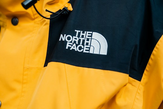 A close-up of a jacket featuring a prominent logo of 'The North Face' embroidered in white on a contrasting black section of fabric. The majority of the jacket is bright yellow with details such as buttons and stitching visible.