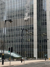 Smoke is billowing out from the windows of a large office building, which is made up of many glass panels. The structure is modern and towering, with several stories visible. In the foreground, there are street lamps and traffic lights, one showing a green light. The street is empty and the day appears overcast.
