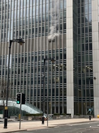 Smoke is billowing out from the windows of a large office building, which is made up of many glass panels. The structure is modern and towering, with several stories visible. In the foreground, there are street lamps and traffic lights, one showing a green light. The street is empty and the day appears overcast.