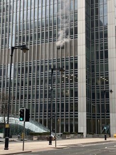 Smoke is billowing out from the windows of a large office building, which is made up of many glass panels. The structure is modern and towering, with several stories visible. In the foreground, there are street lamps and traffic lights, one showing a green light. The street is empty and the day appears overcast.