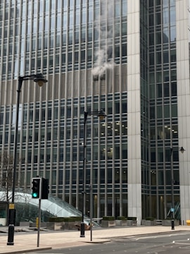 Smoke is billowing out from the windows of a large office building, which is made up of many glass panels. The structure is modern and towering, with several stories visible. In the foreground, there are street lamps and traffic lights, one showing a green light. The street is empty and the day appears overcast.
