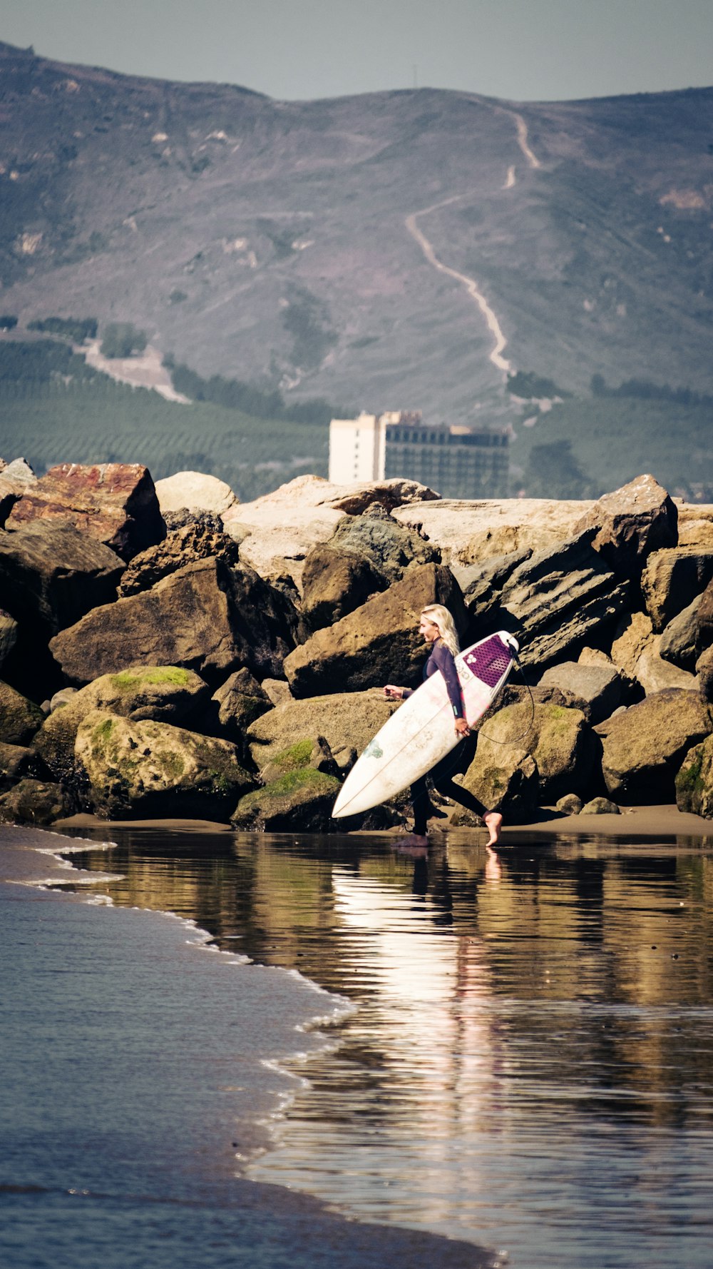 분홍색 긴팔 셔츠와 흰 바지를 입은 여자가 바위 해안에 서 있는 흰색 서핑보드를 들고 있다