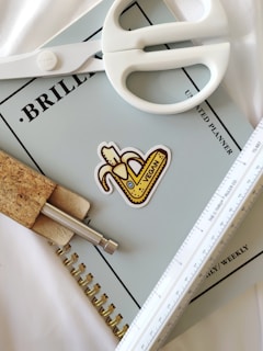 A pair of white scissors is placed on top of a light blue planner titled 'Brilliant' with additional text indicating it's an unlimited planner. A sticker featuring a banana and the word 'vegan' is also on the planner. A cork pen and a white ruler are positioned alongside.