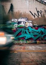 Graffiti covers a concrete wall with colorful designs and vibrant lettering, including a combination of abstract and stylized words. A blurry car with headlights on zooms past in the foreground, adding a sense of motion and urban dynamism.