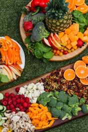 A colorful arrangement of fruits, vegetables, nuts, and seeds displayed on wooden platters over grass. There is a variety of produce such as pineapple, oranges, apples, strawberries, raspberries, broccoli, cauliflower, sliced cucumber, bell pepper, and lettuce.