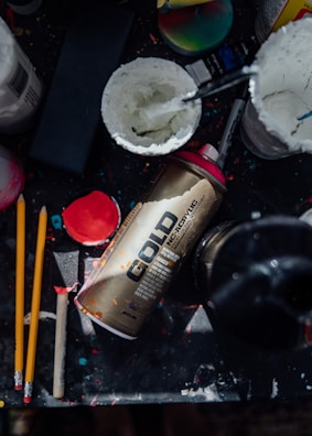 An arrangement of art supplies including a gold spray paint can labeled 'GOLD', two yellow pencils, a black sponge brush, and containers filled with white paint. The background is covered in colorful paint splatters.