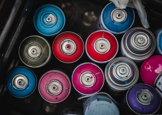 A collection of colorful spray paint cans arranged in a container, showcasing various shades including blue, red, and pink. The cans are seen from above, highlighting their metal nozzles and circular tops.